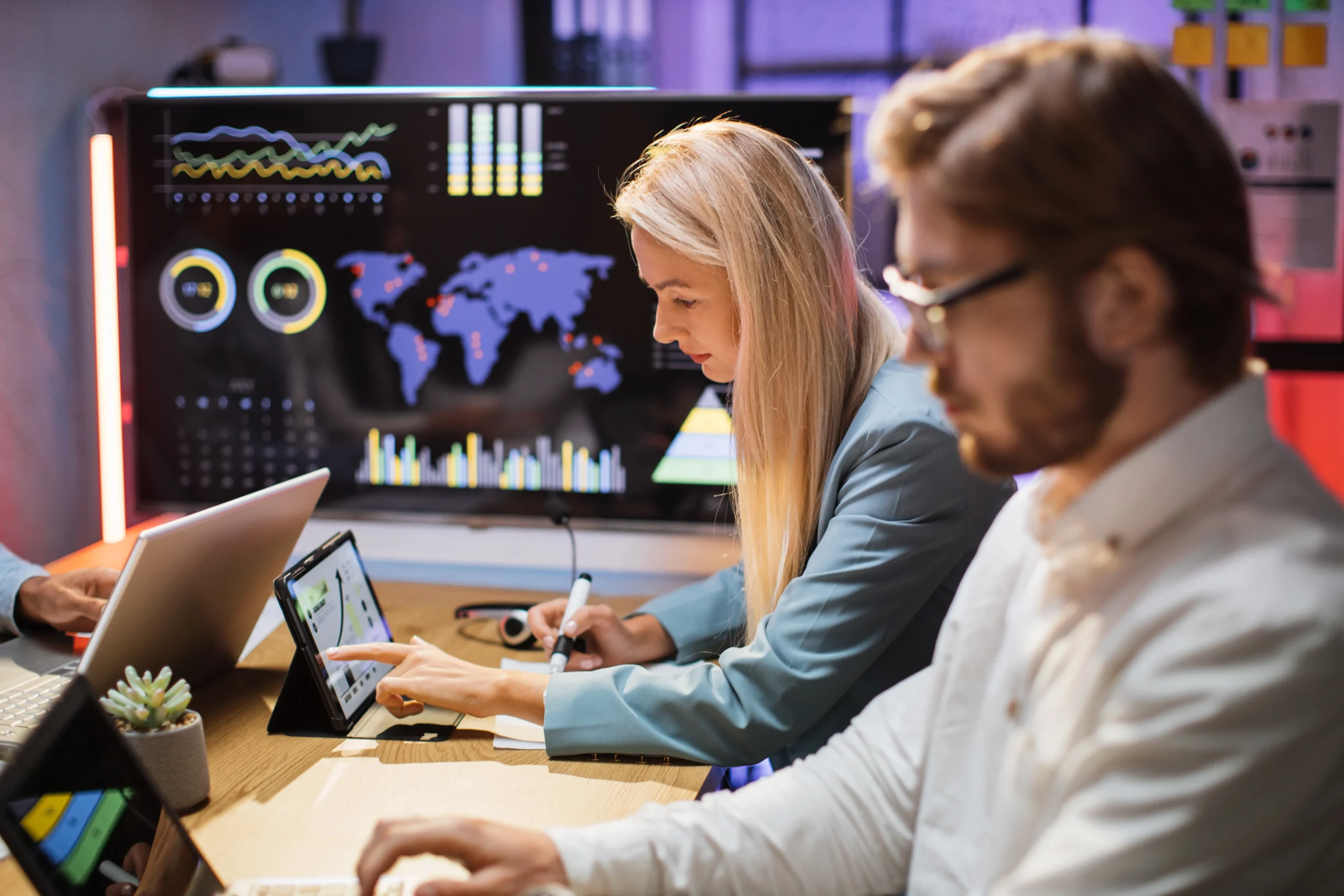 Two people work at computers with charts and world maps displayed on large monitors in the background, analyzing data in a modern office setting.