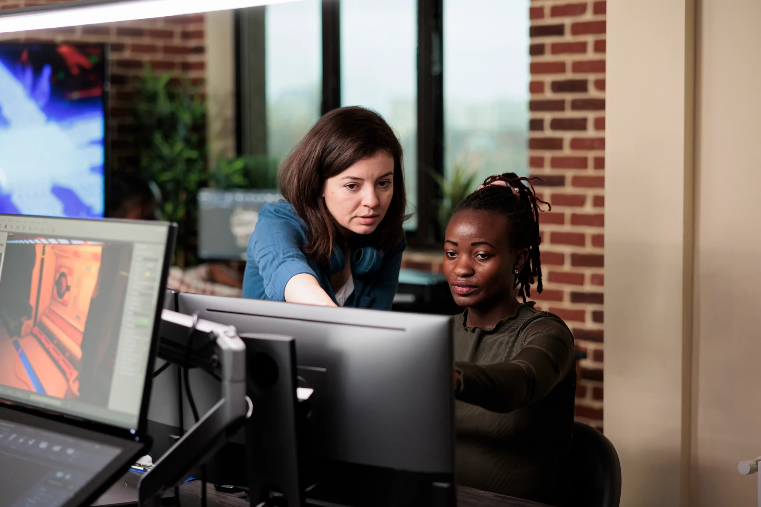 Two women work together at a computer in an office with brick walls; one is pointing at the monitor while the other looks at the screen.