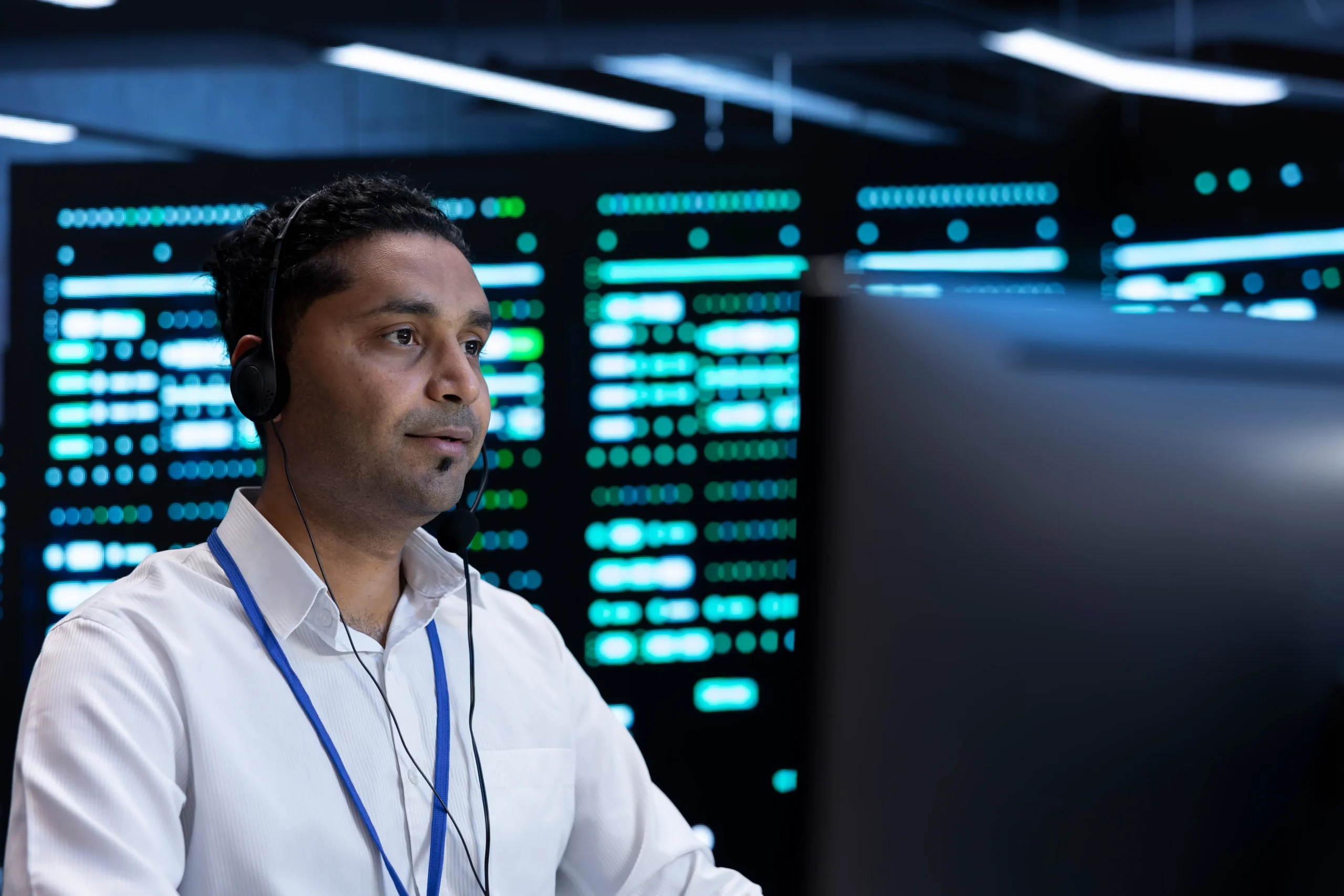 A man wearing a headset and ID badge sits at a computer in a modern server room with illuminated monitors and equipment in the background.