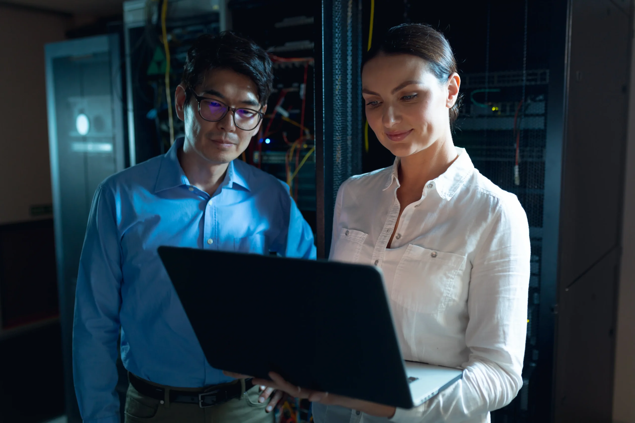 Two people standing in a server room, looking at a laptop together, with server racks and network cables visible in the background.