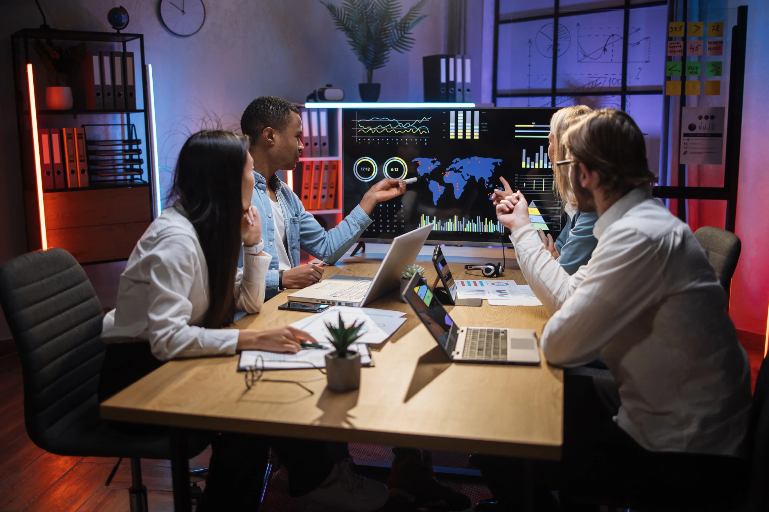 Four people sit at a conference table with laptops, reviewing data and graphs displayed on a large screen in a modern office setting.