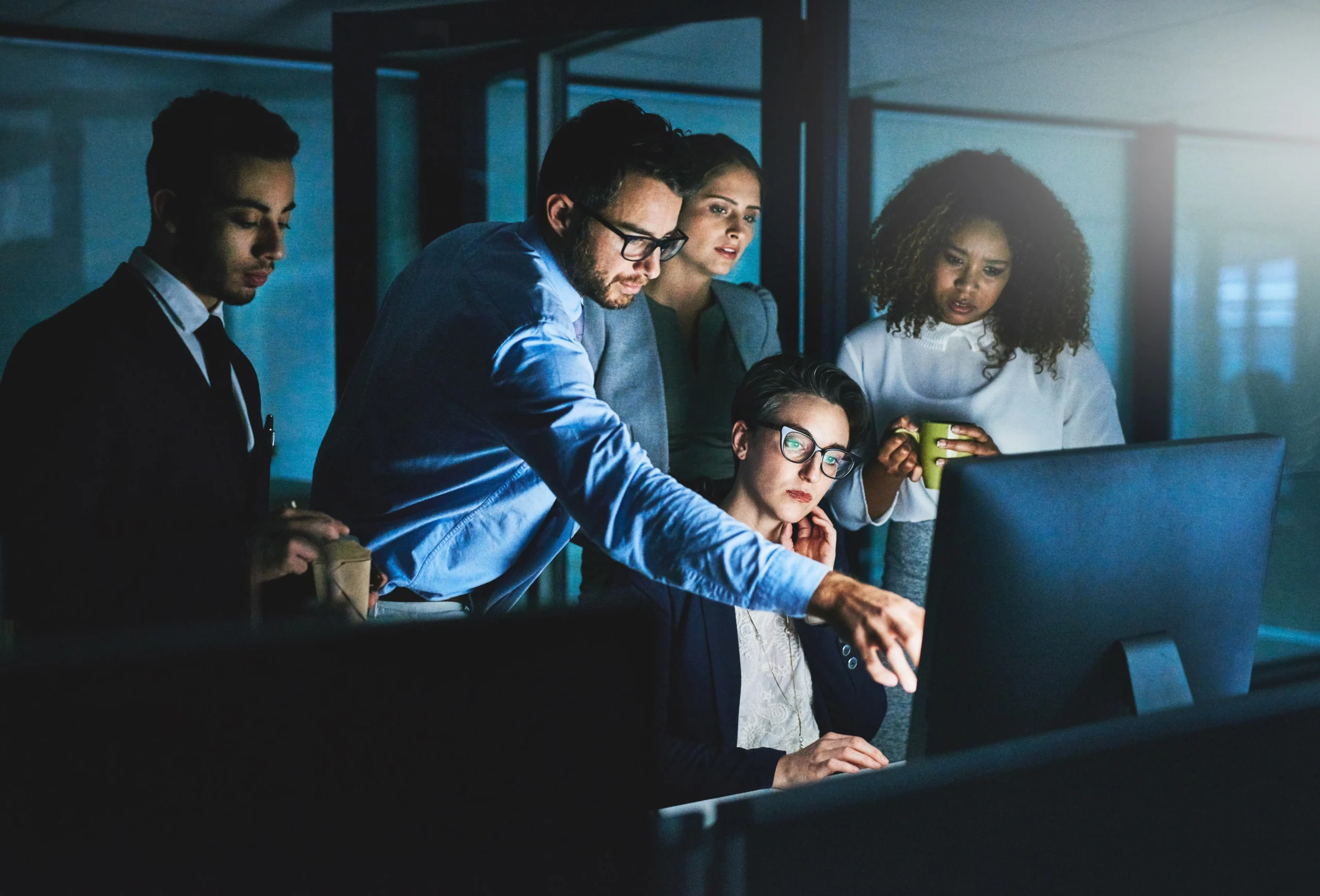 Five people gather around a computer monitor in an office at night, focusing intently as one person points to the screen and others observe or hold drinks.