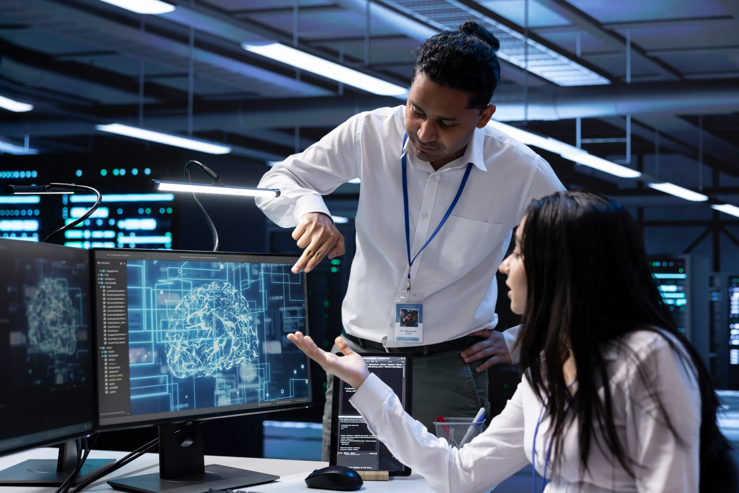 Two people in an office discuss data on a computer screen displaying a digital brain and technical diagrams, suggesting a technology or AI project.