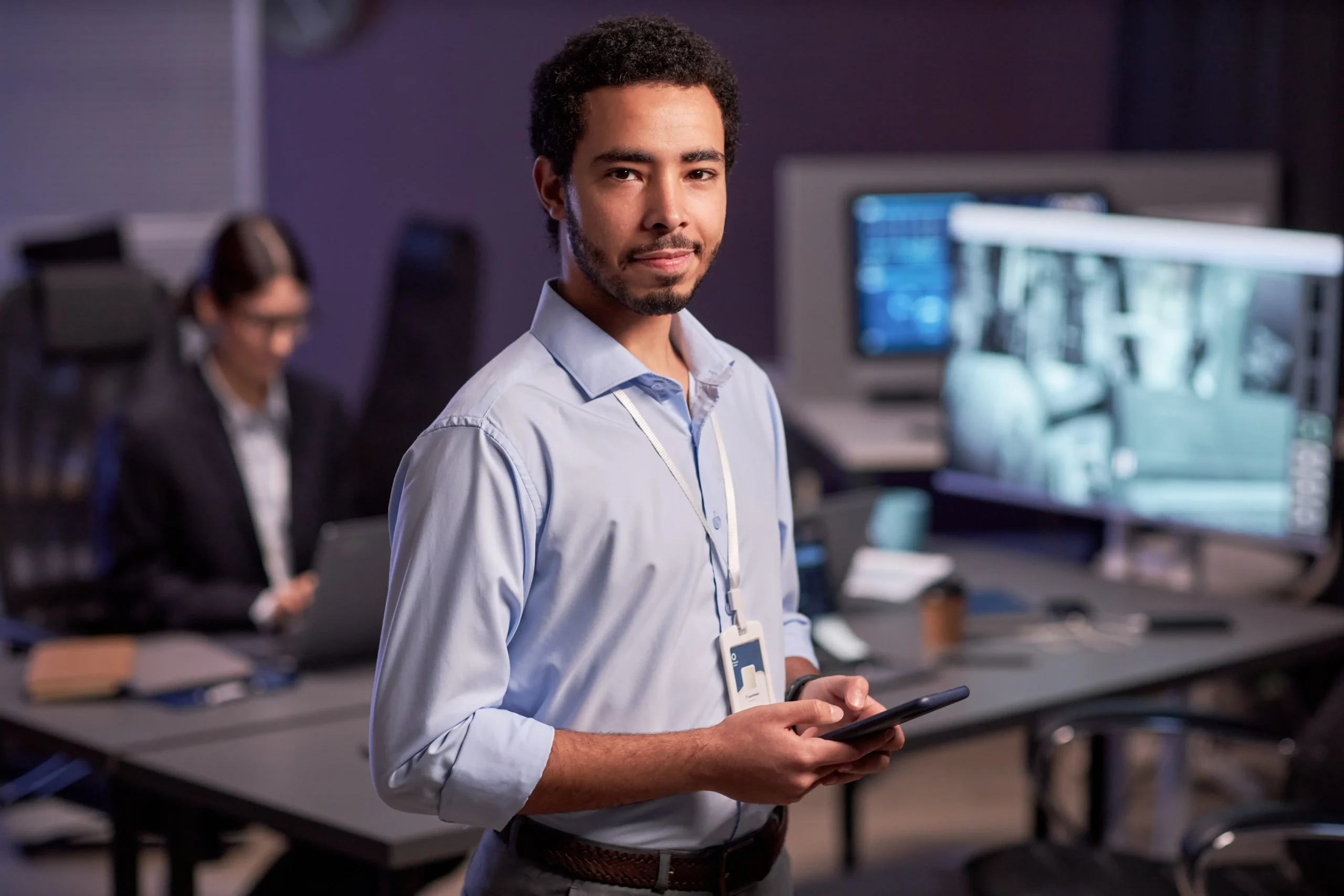 A man in a blue shirt holding a tablet stands in an office with computer monitors, while another person works at a desk in the background.