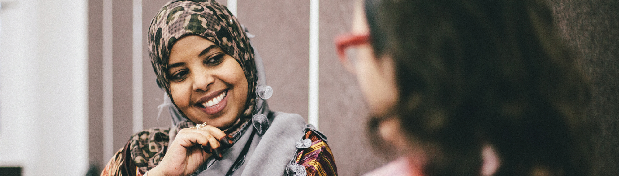 A woman wearing a patterned headscarf smiles and listens to another person with long hair and red glasses in a conversation.
