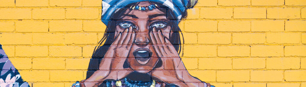 A mural on a yellow brick wall depicts a woman wearing a headscarf, holding her hands to her mouth as if shouting or calling out.