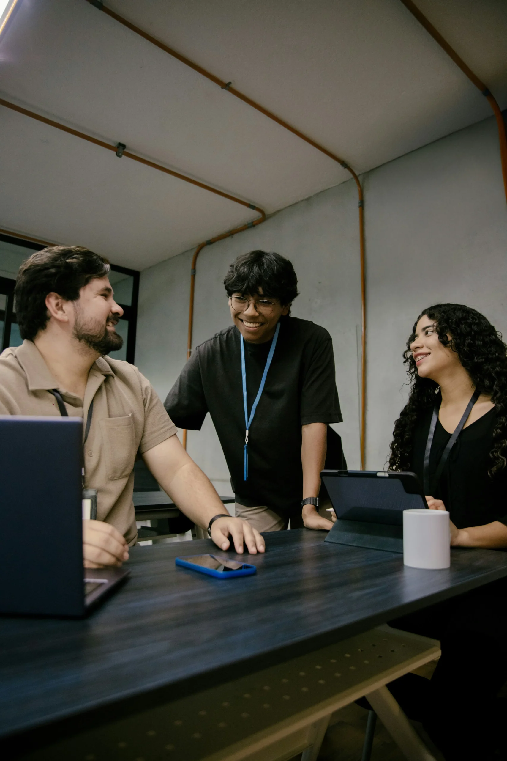 Three people are having a discussion around a table with laptops, a tablet, and a coffee mug in a modern office setting.