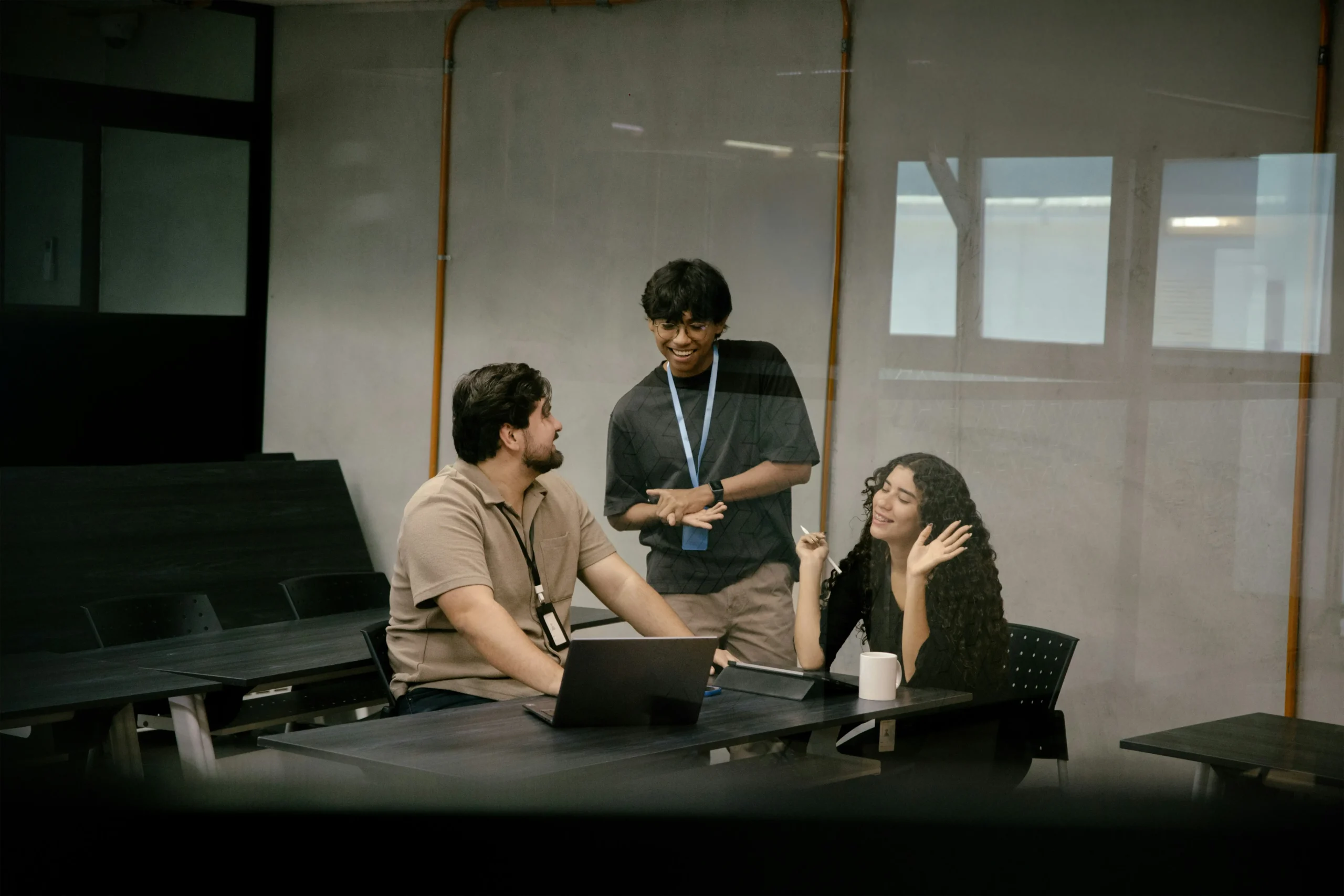 Three people in an office setting are having a discussion around a table with a laptop, viewed through a glass window.
