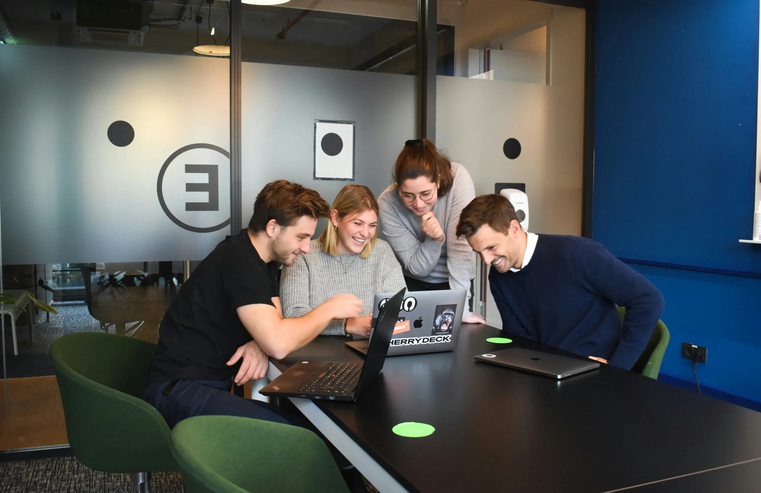 Four people sit and stand around a table, looking at a laptop screen and smiling in a modern office meeting room.