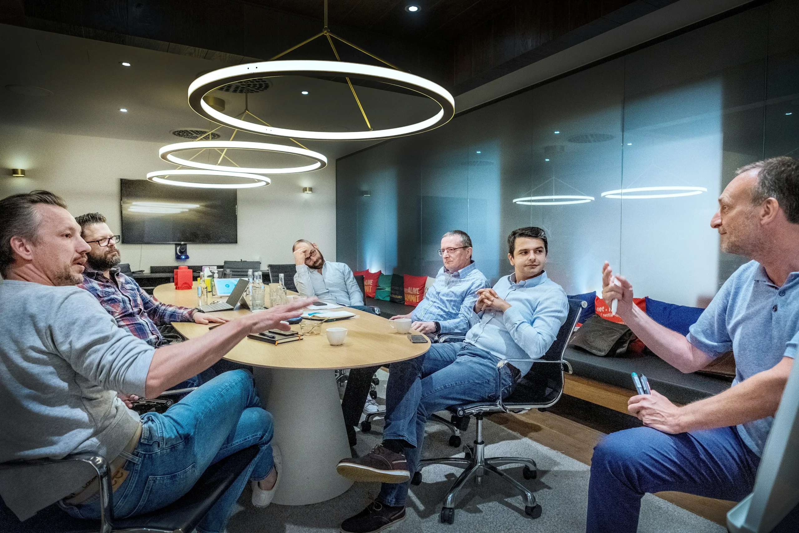 Six men sit around a conference table in a modern office, engaged in discussion. Three circular ceiling lights hang above, and notepads, drinks, and cups are on the table.
