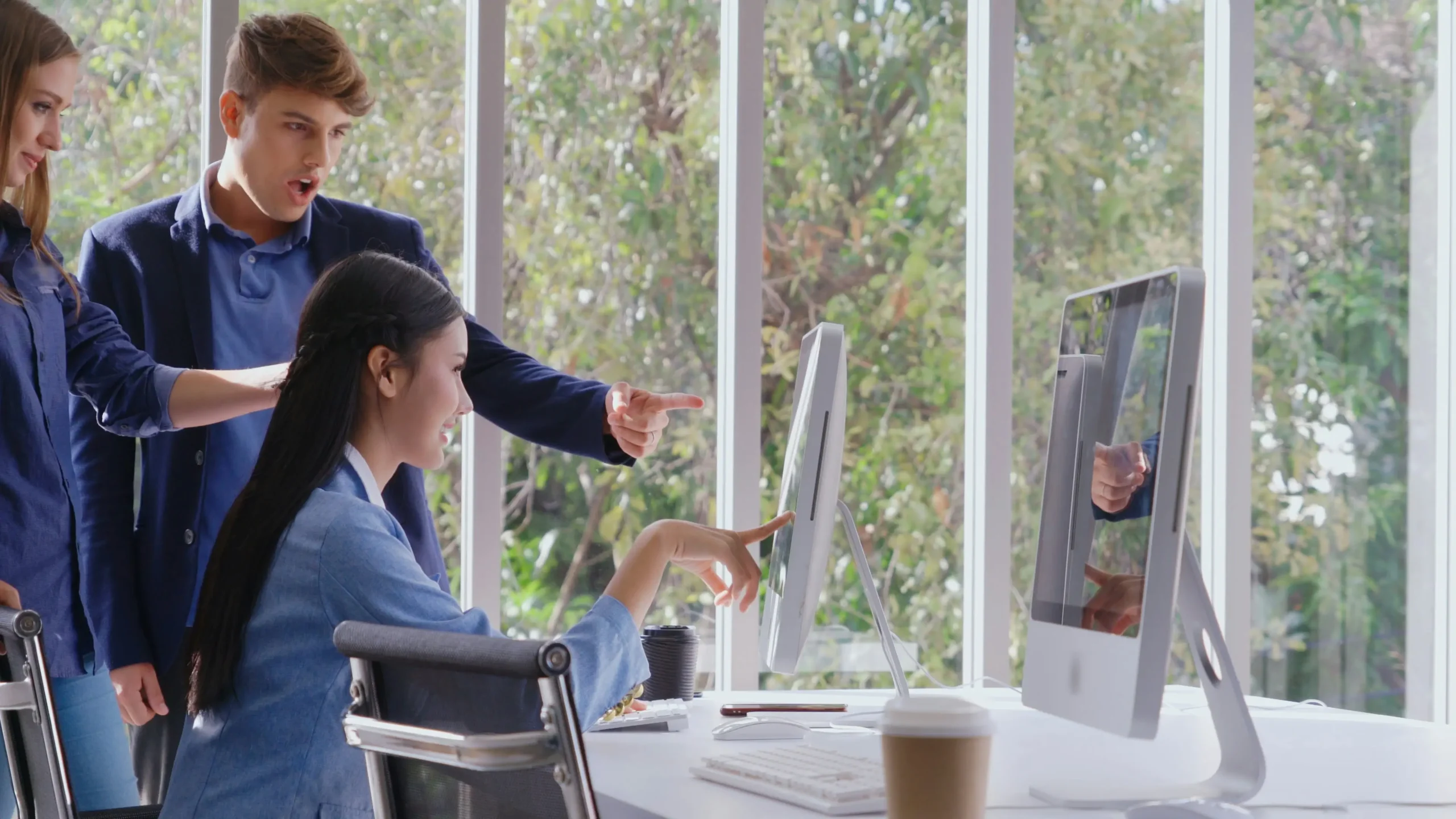 Three people in business attire discuss something on a computer screen in a bright office with large windows and trees visible outside.