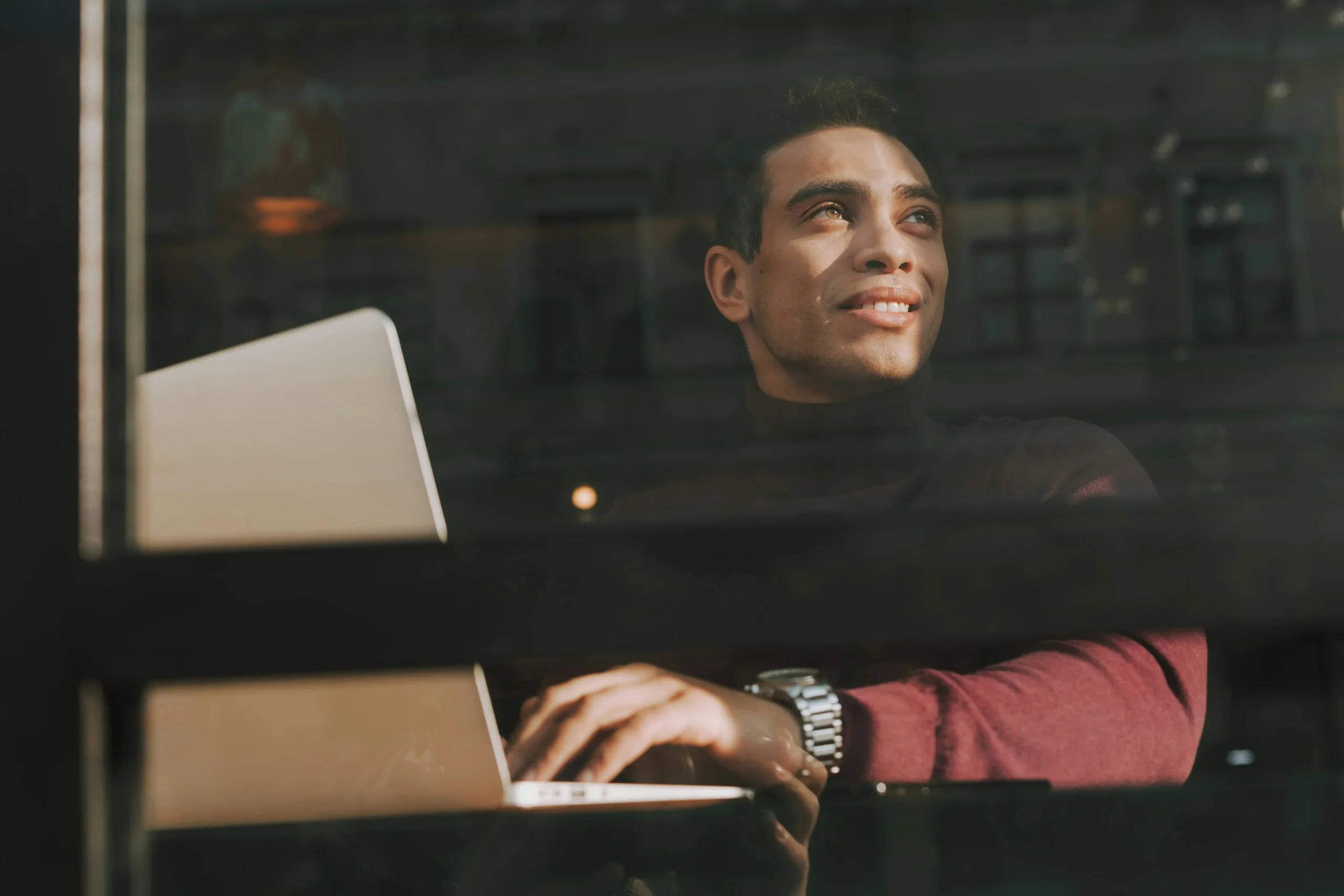 A man sits at a table with a laptop, looking out of a window with a slight smile, his hands resting on the keyboard.