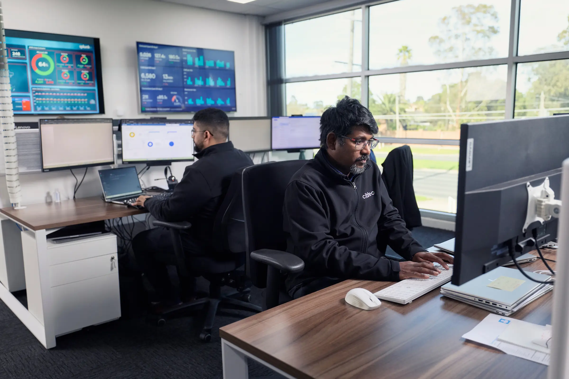Two men are working at desks with multiple computer monitors in a modern office with large windows and digital dashboards on the wall.
