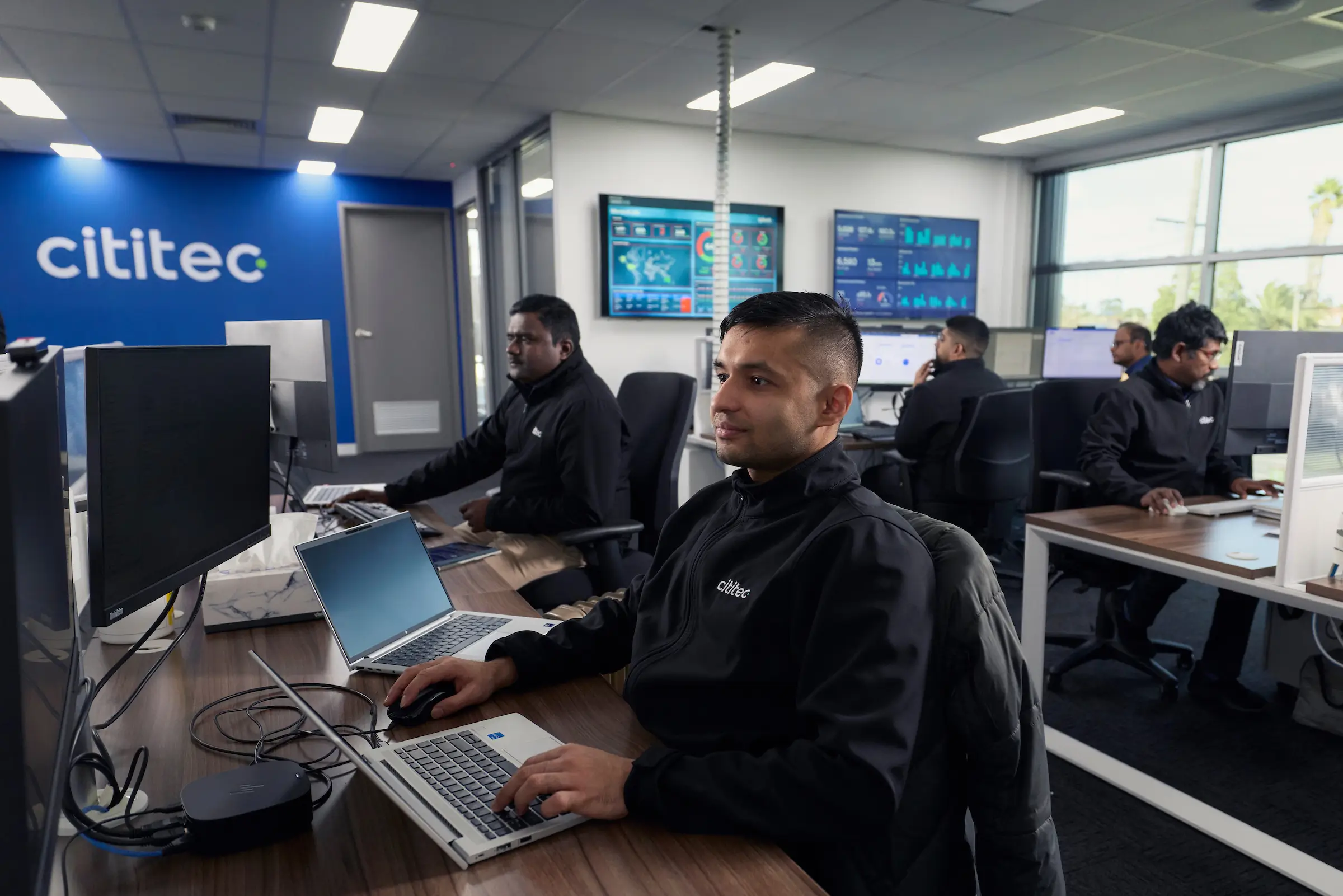 Several people work at computers in a modern office with “cititec” branding on the wall and multiple monitors displaying data.