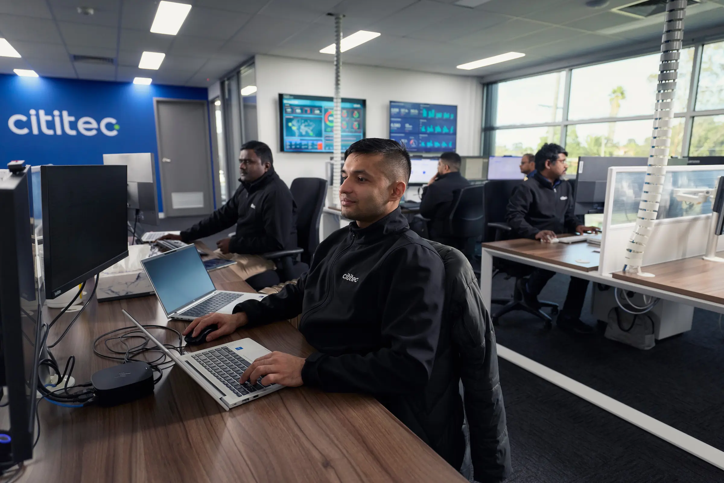 Five people work at computers in a modern office space with "cititec" branding visible on the wall.