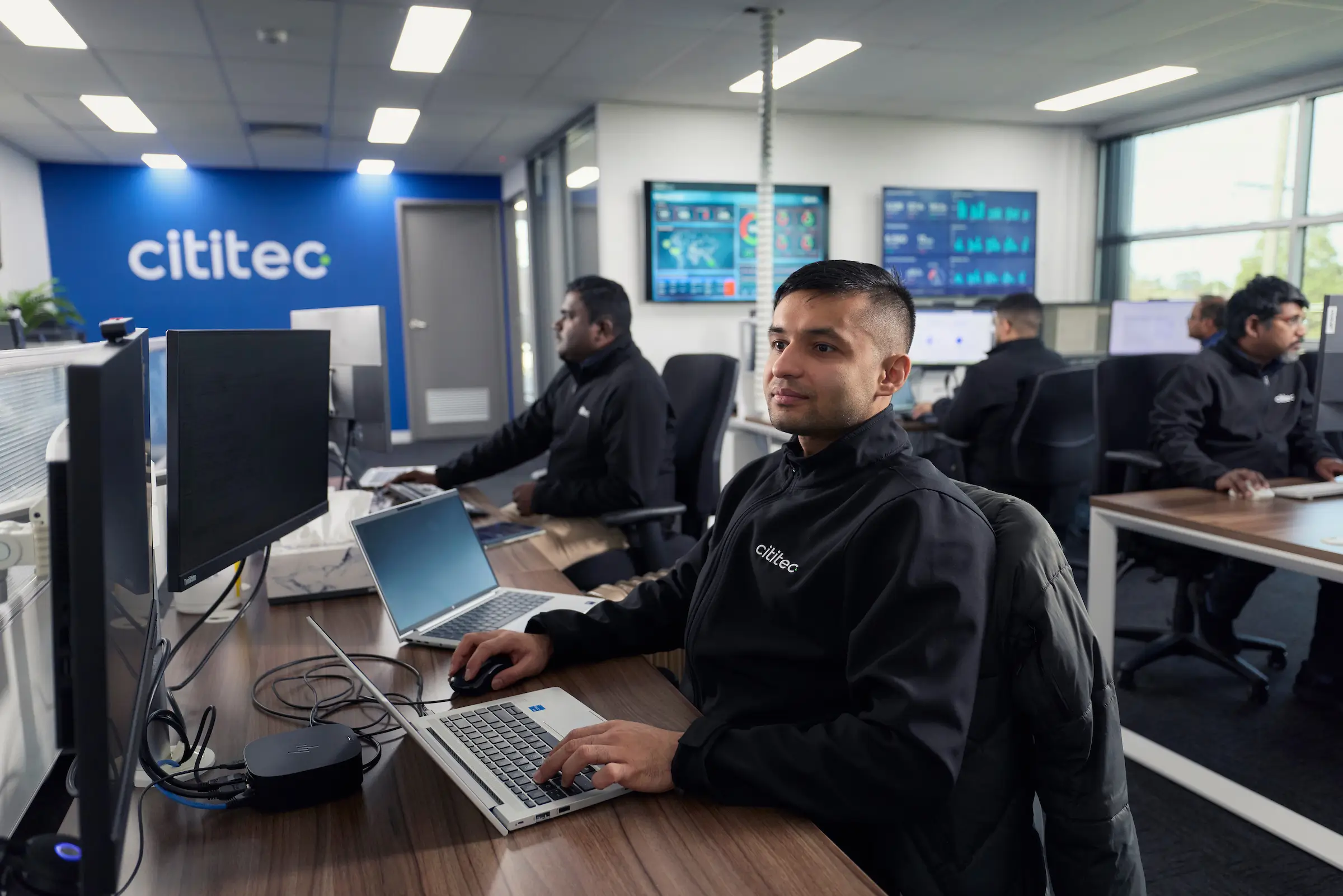 Several people work at desks with computers in a modern office. One person in the foreground looks toward the camera. Cititec branding is visible on a wall.