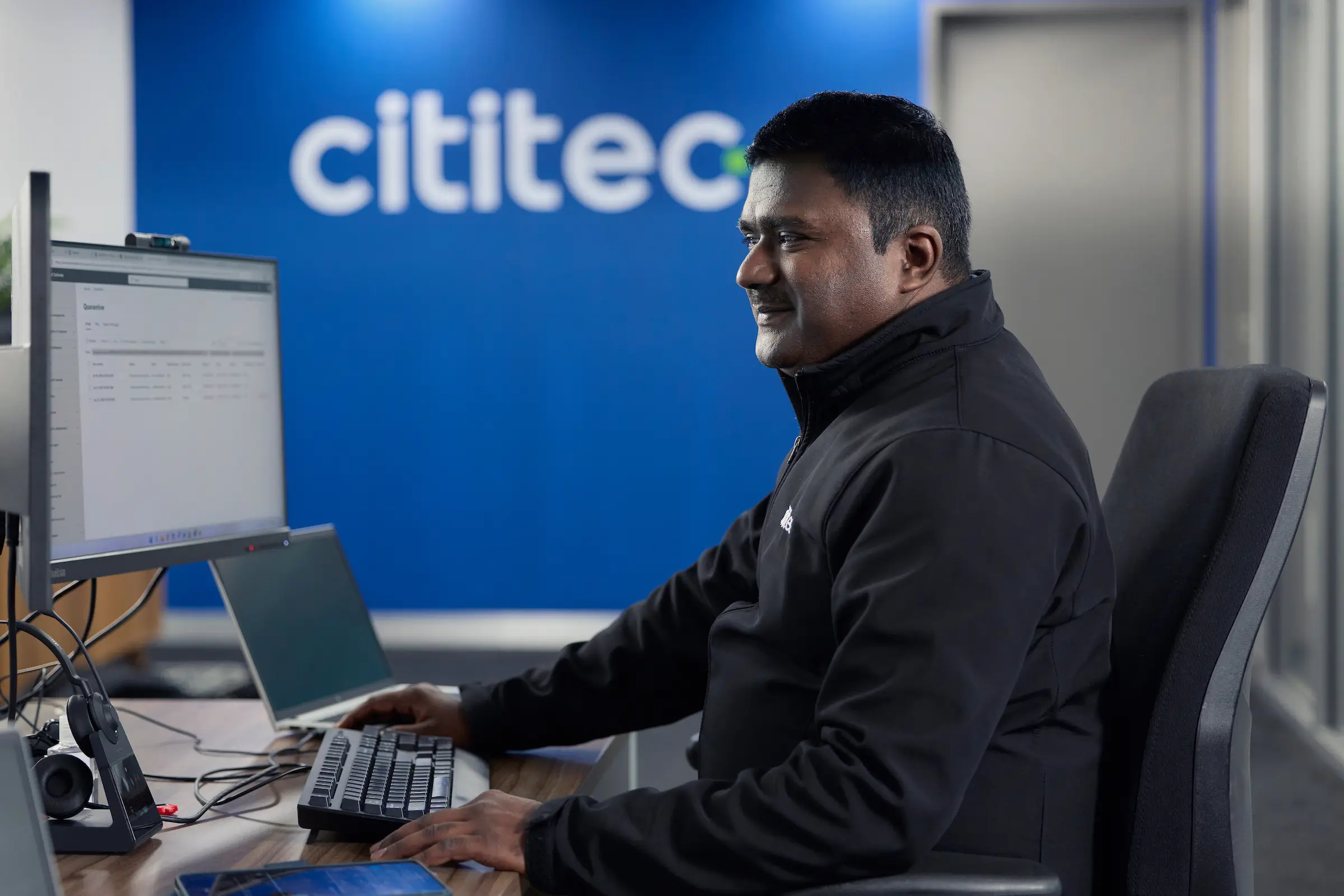 A man sits at a desk working on a computer in an office; the wall behind him displays the Cititec logo.