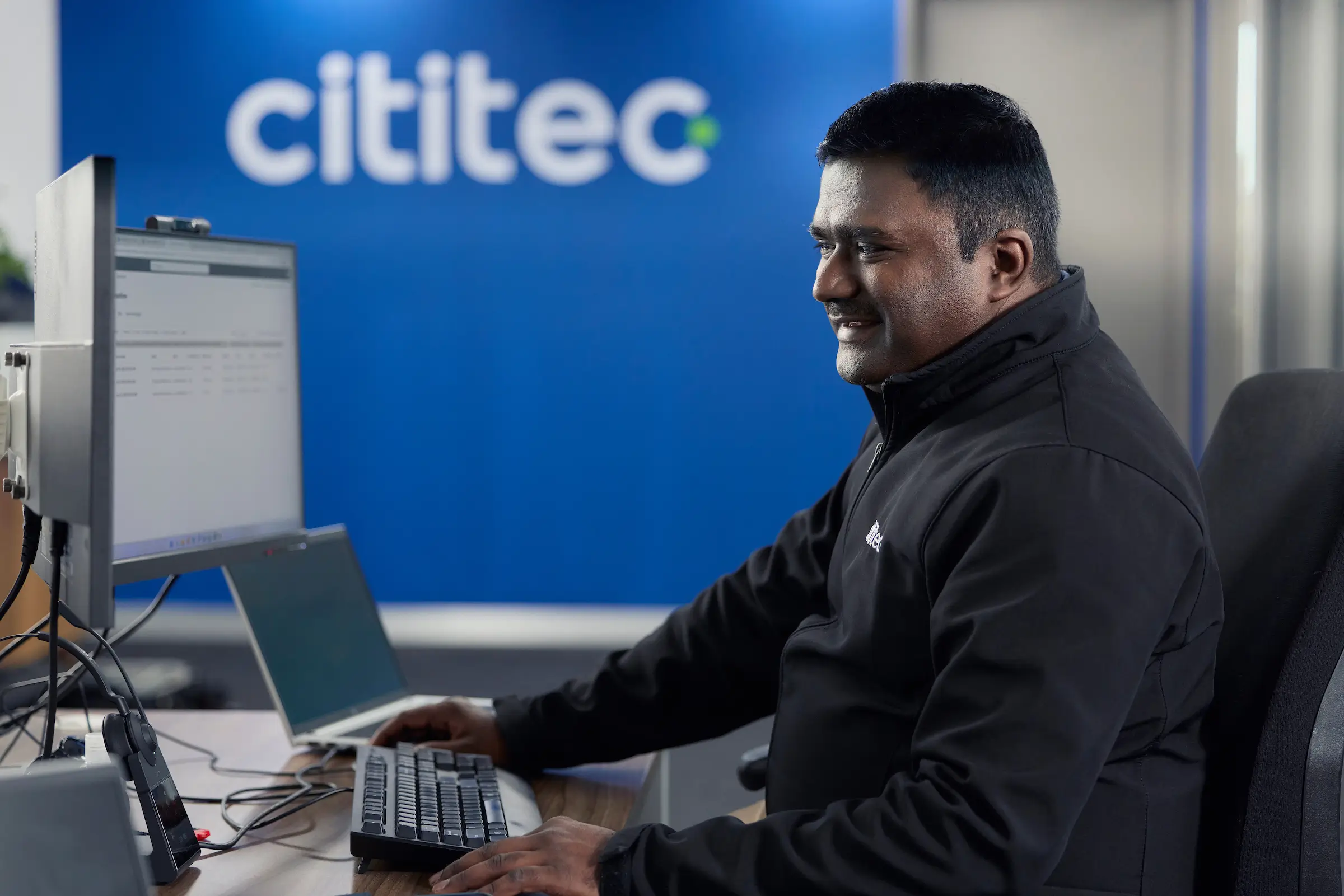 A man working at a computer in an office, with the Cititec logo visible on a blue wall in the background.