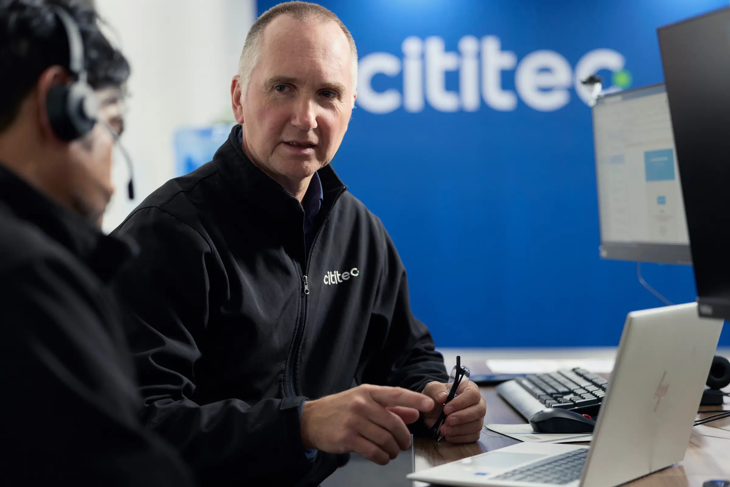 Two men in Cititec-branded jackets sit at a desk with computers, engaged in conversation in an office with a Cititec sign in the background.