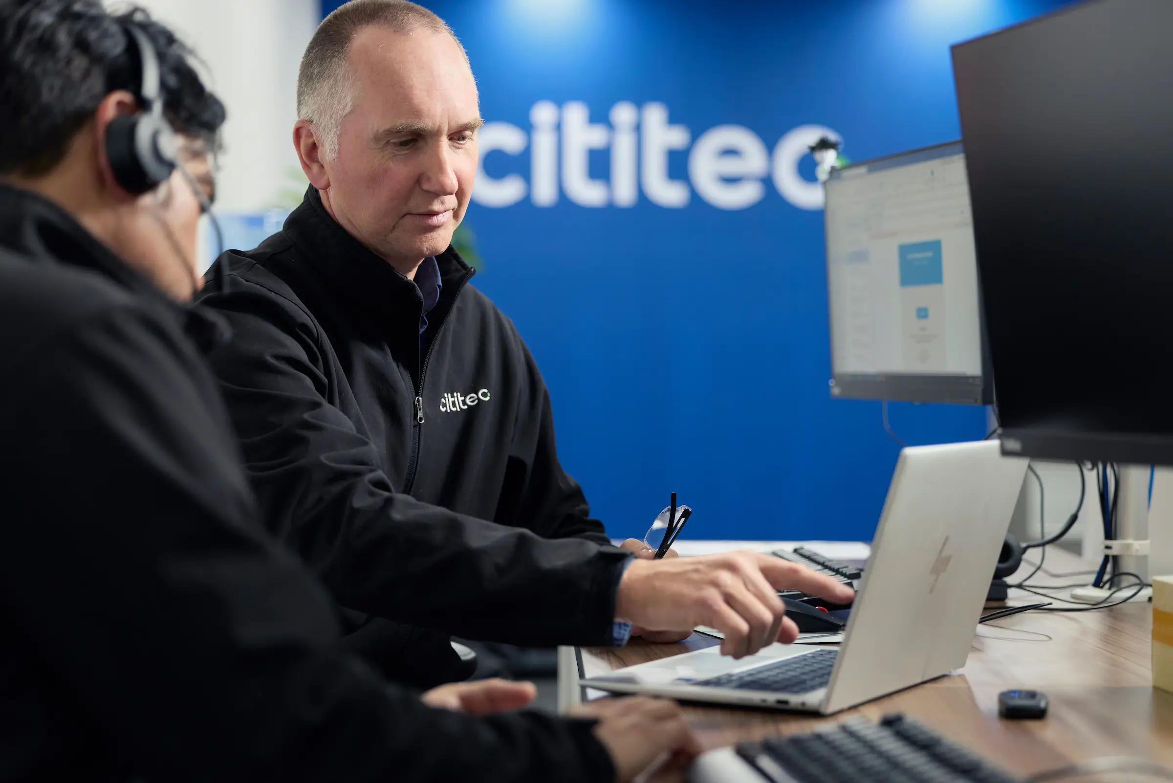 Two men working at computers in an office, one pointing at a laptop screen, with "cititec" logo visible on the wall in the background.
