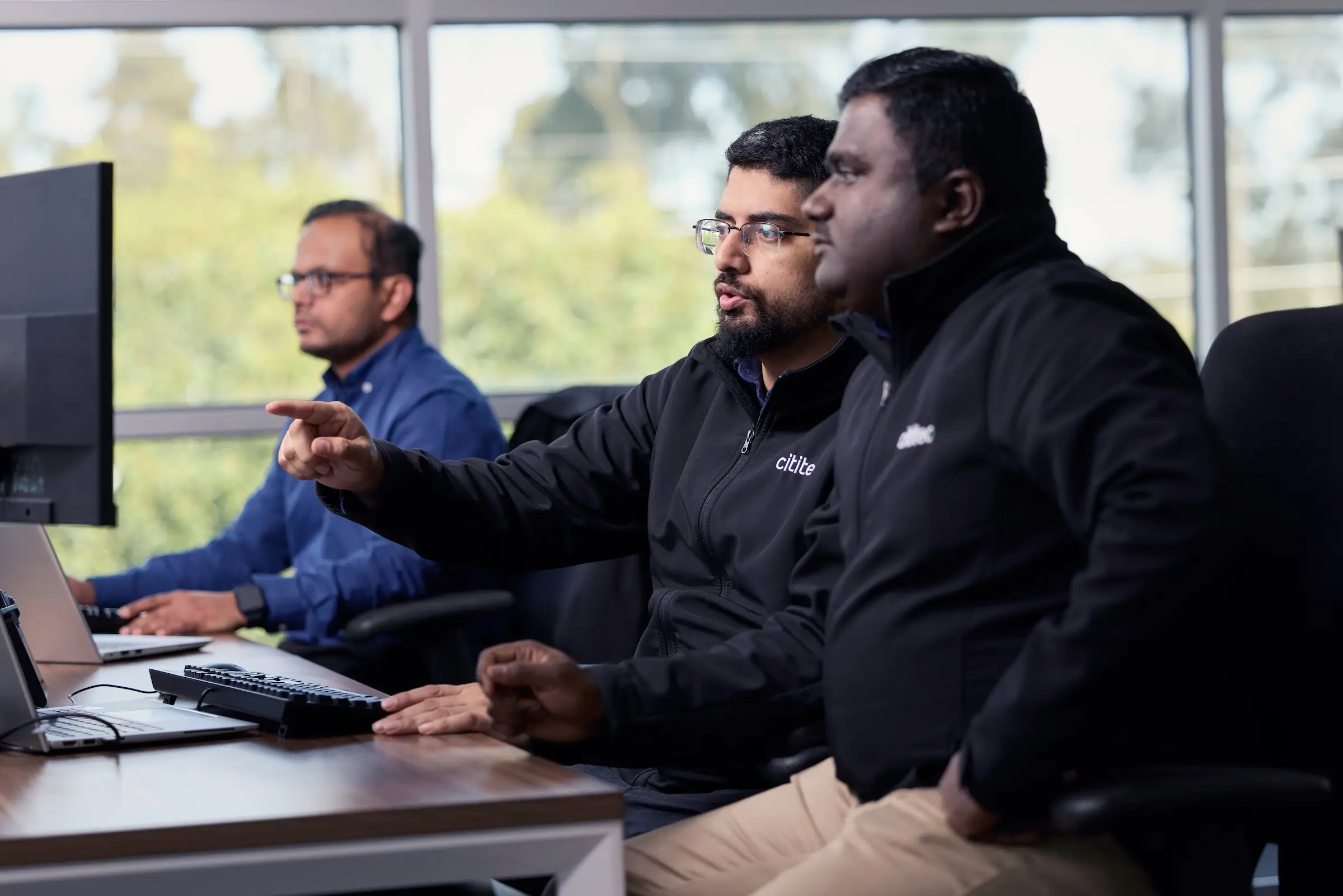 Three men sit at desks working on computers; two are discussing something on a screen while the third focuses on his own monitor in the background.