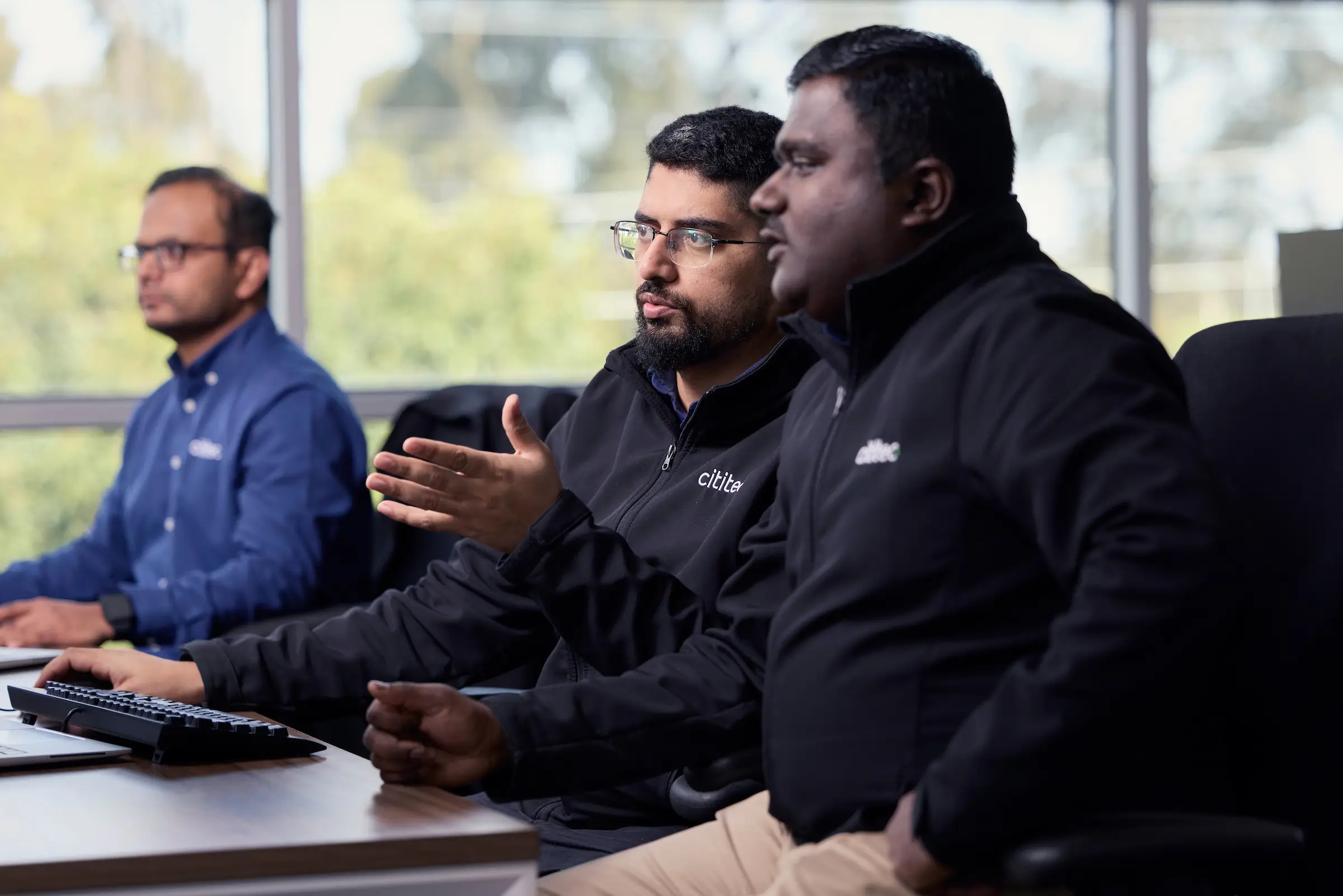 Three men sit at desks working on computers. Two are in black jackets engaged in discussion, while the third man in a blue shirt focuses on his screen. Large windows are in the background.