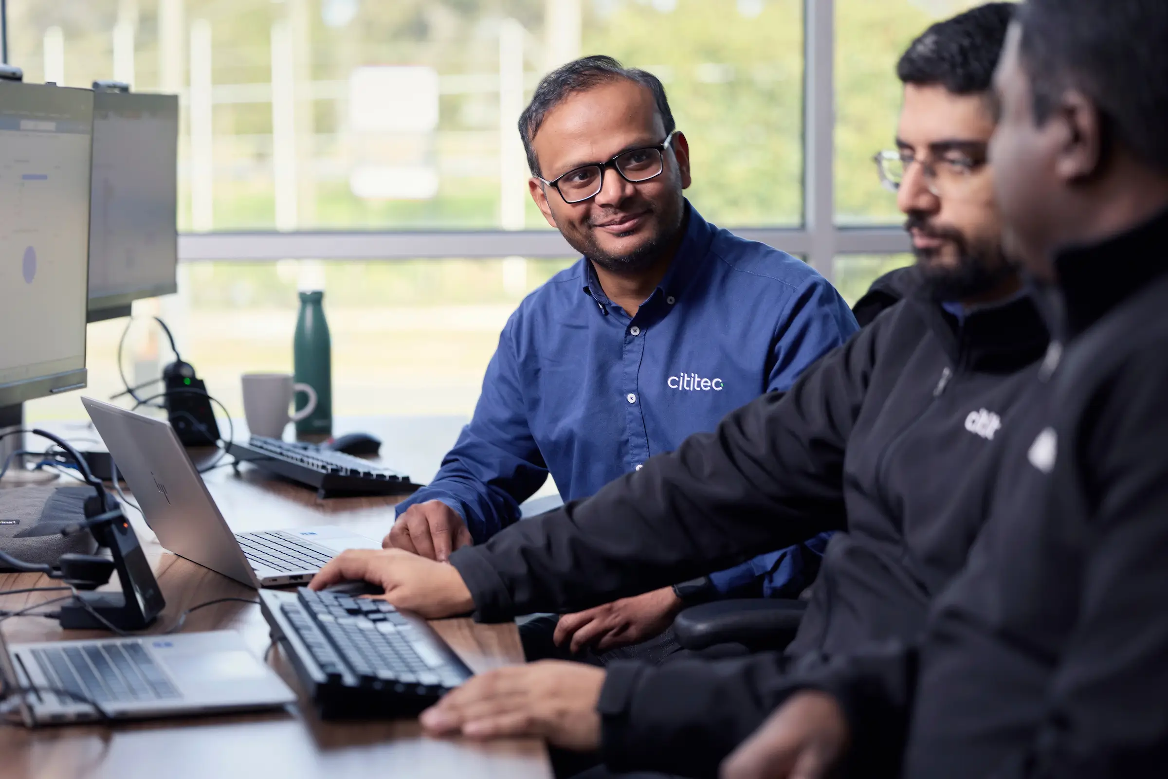 Three men sit at a desk with computers, working and conversing in an office setting. One is wearing a blue shirt with a "citec" logo, the others wear black jackets.