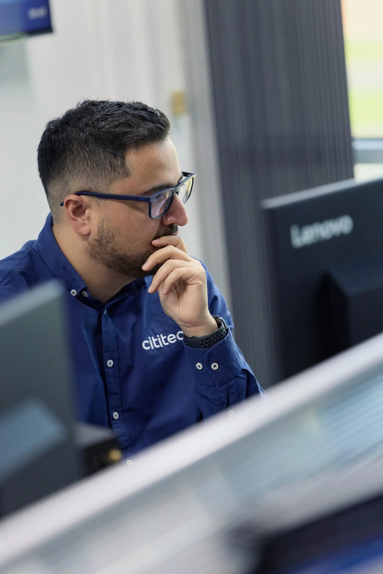 A man wearing glasses and a blue shirt labeled "clinitec" sits at a desk, looking at a computer monitor and resting his hand on his chin.
