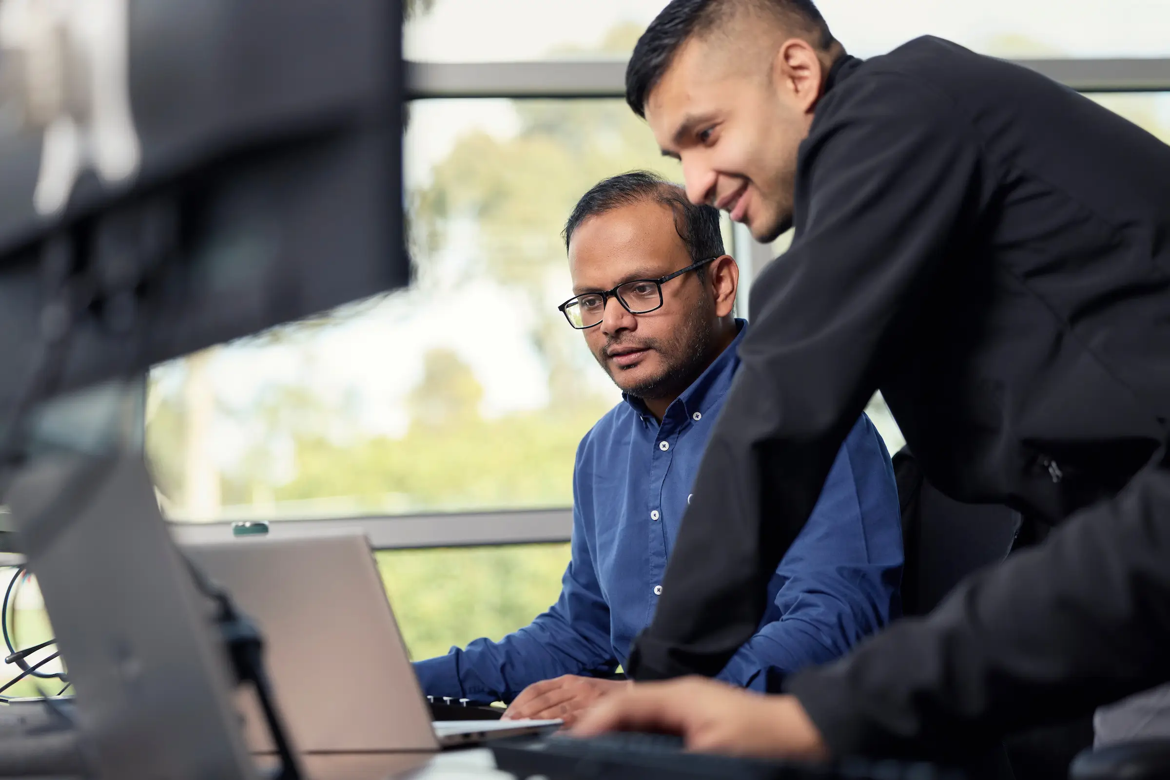 Two men working at computers in an office, one sitting and looking at a laptop screen, the other standing and pointing at a monitor.