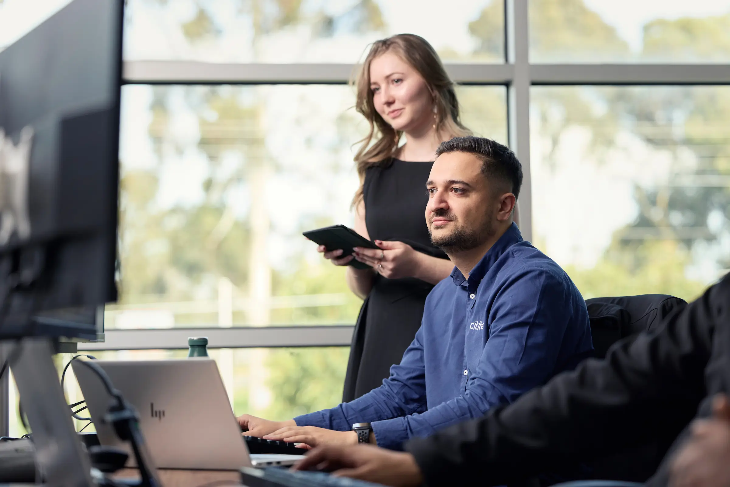 A man sits at a desk working on a computer while a woman stands behind him holding a tablet in an office with large windows.
