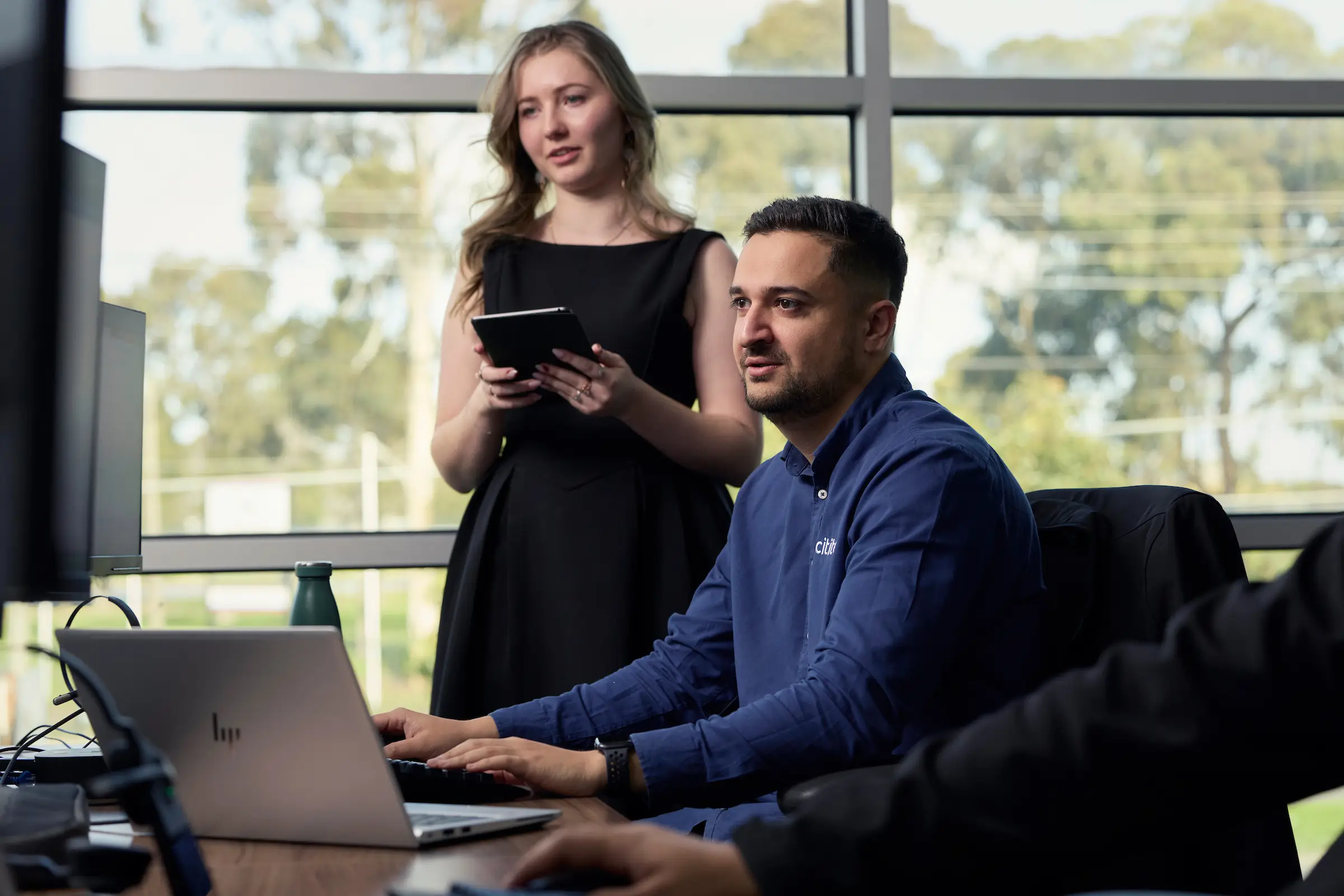 A man works at a computer while a woman stands beside him holding a tablet. Both are focused on the screen in an office setting with large windows.