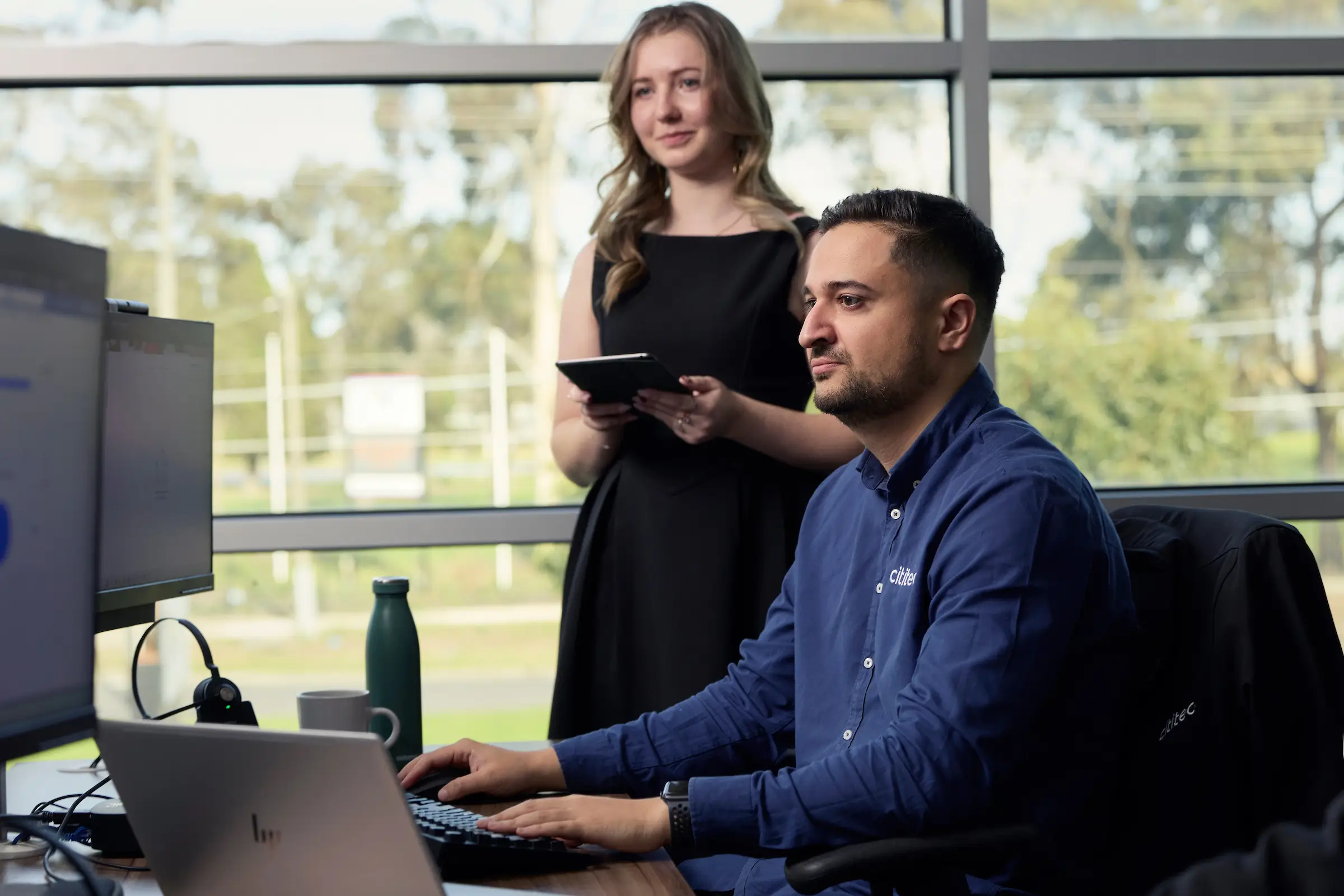 A man works at a computer while a woman stands behind him holding a tablet; they are in an office with large windows in the background.
