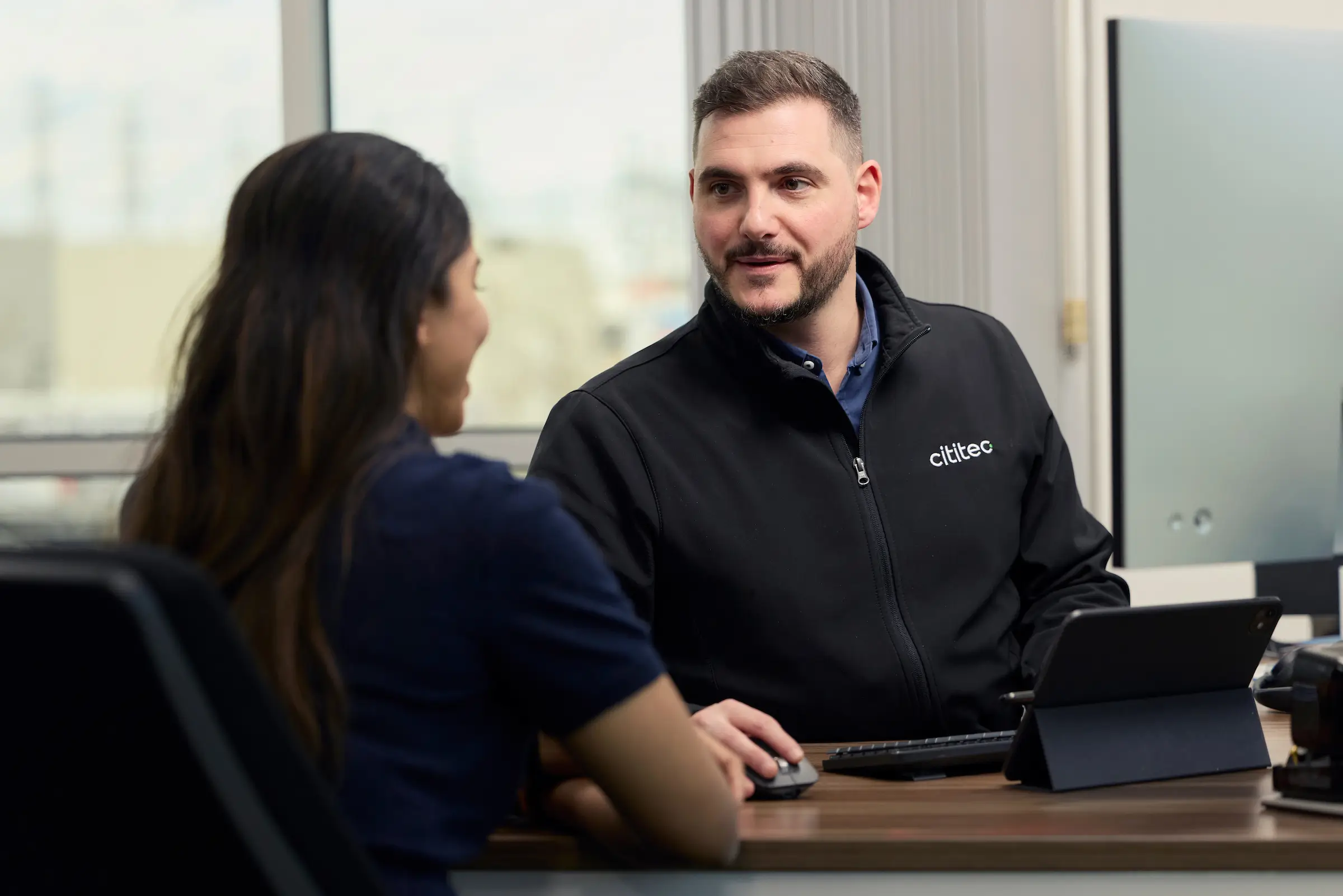 A man in a Citibee-branded jacket sits at a desk with a computer, talking to a woman seated across from him in an office setting.