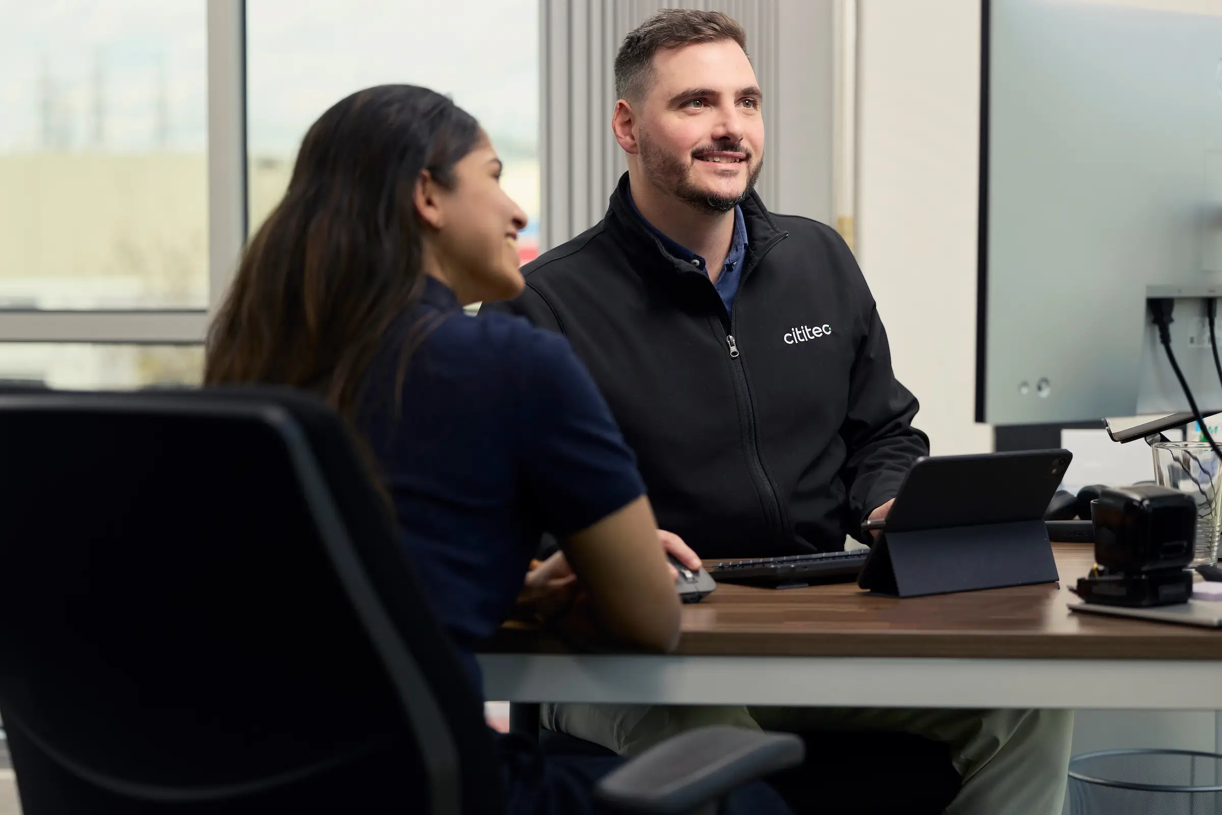 Two people sit at an office desk, one facing a computer monitor and the other using a tablet, engaged in conversation.