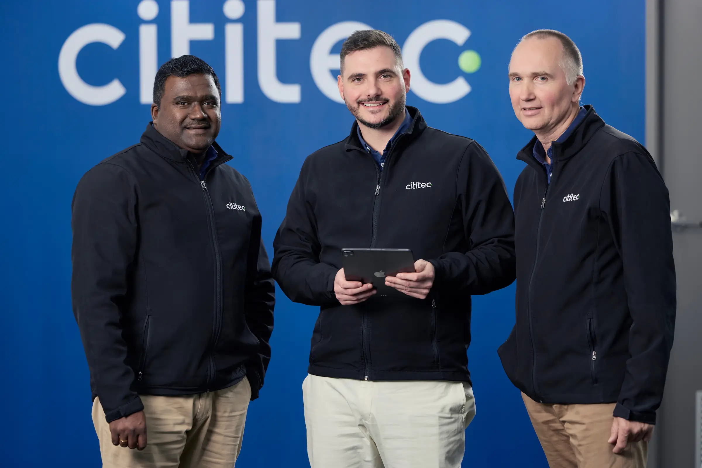 Three men wearing matching black Cititec jackets stand in front of a blue wall with the Cititec logo; one holds a tablet.