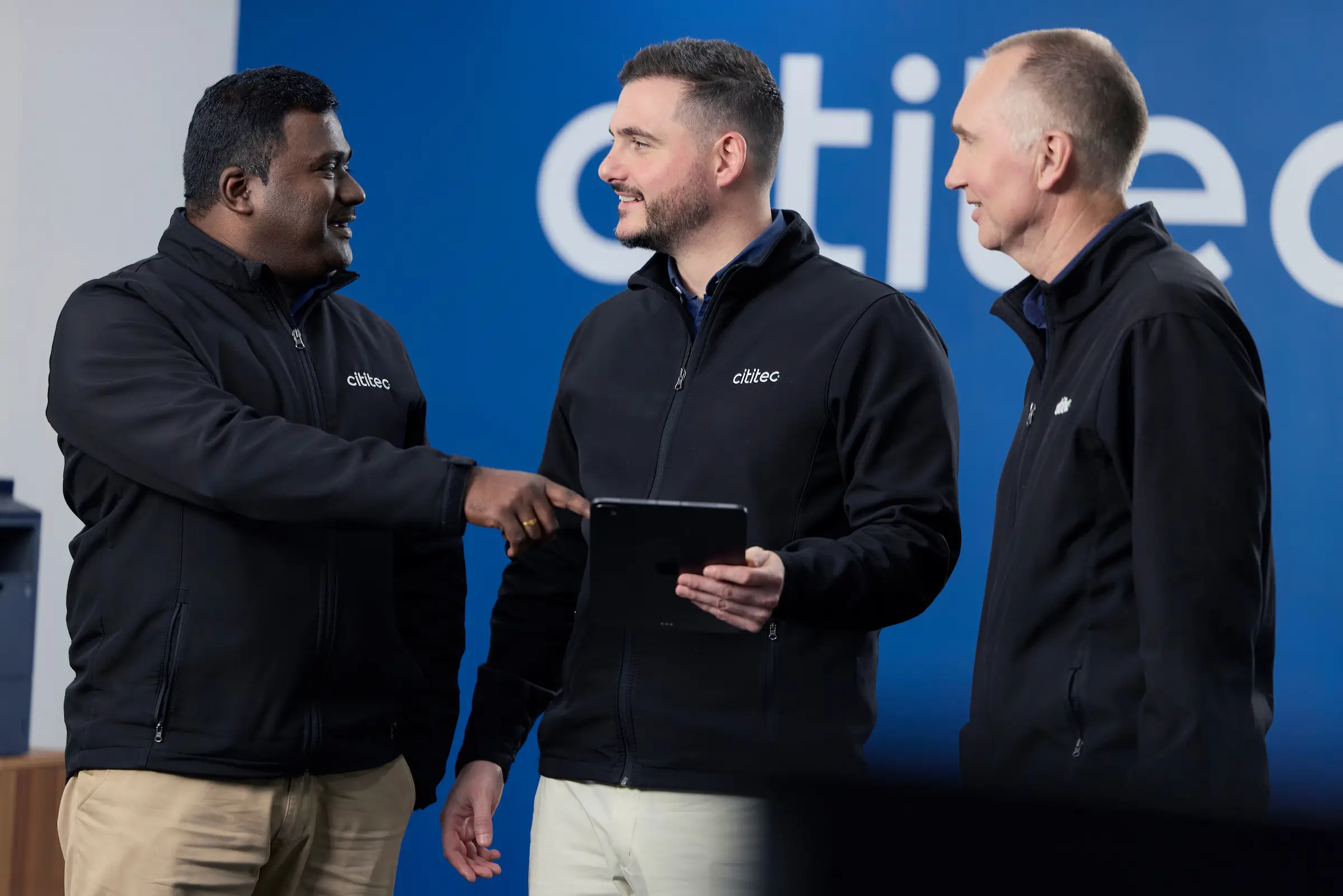 Three men in black jackets stand together indoors, with one holding a tablet. They appear to be having a discussion in front of a blue wall with partial white text.