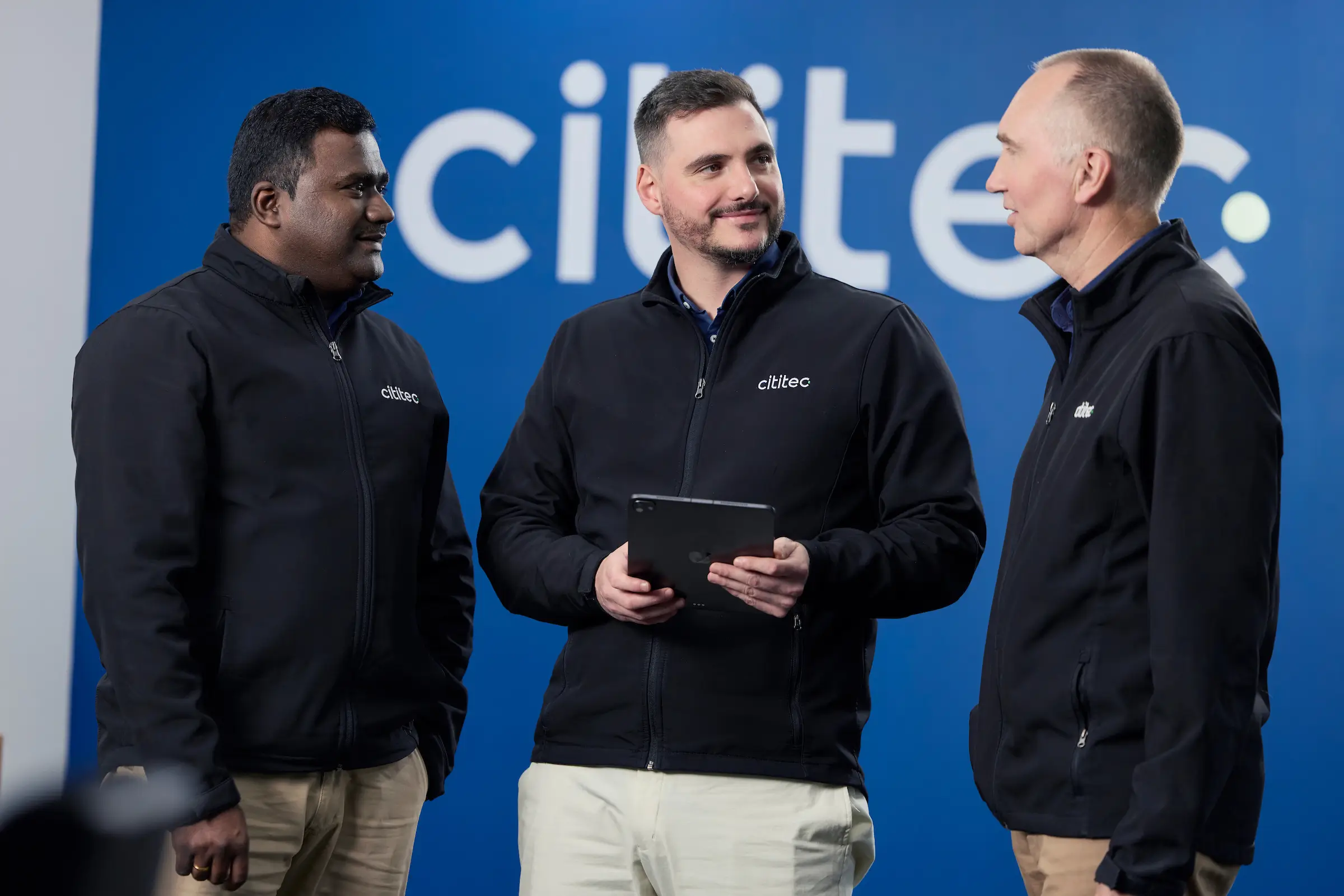 Three men wearing black jackets stand in front of a blue background with "cilitec" logo; one holds a tablet while they engage in conversation.
