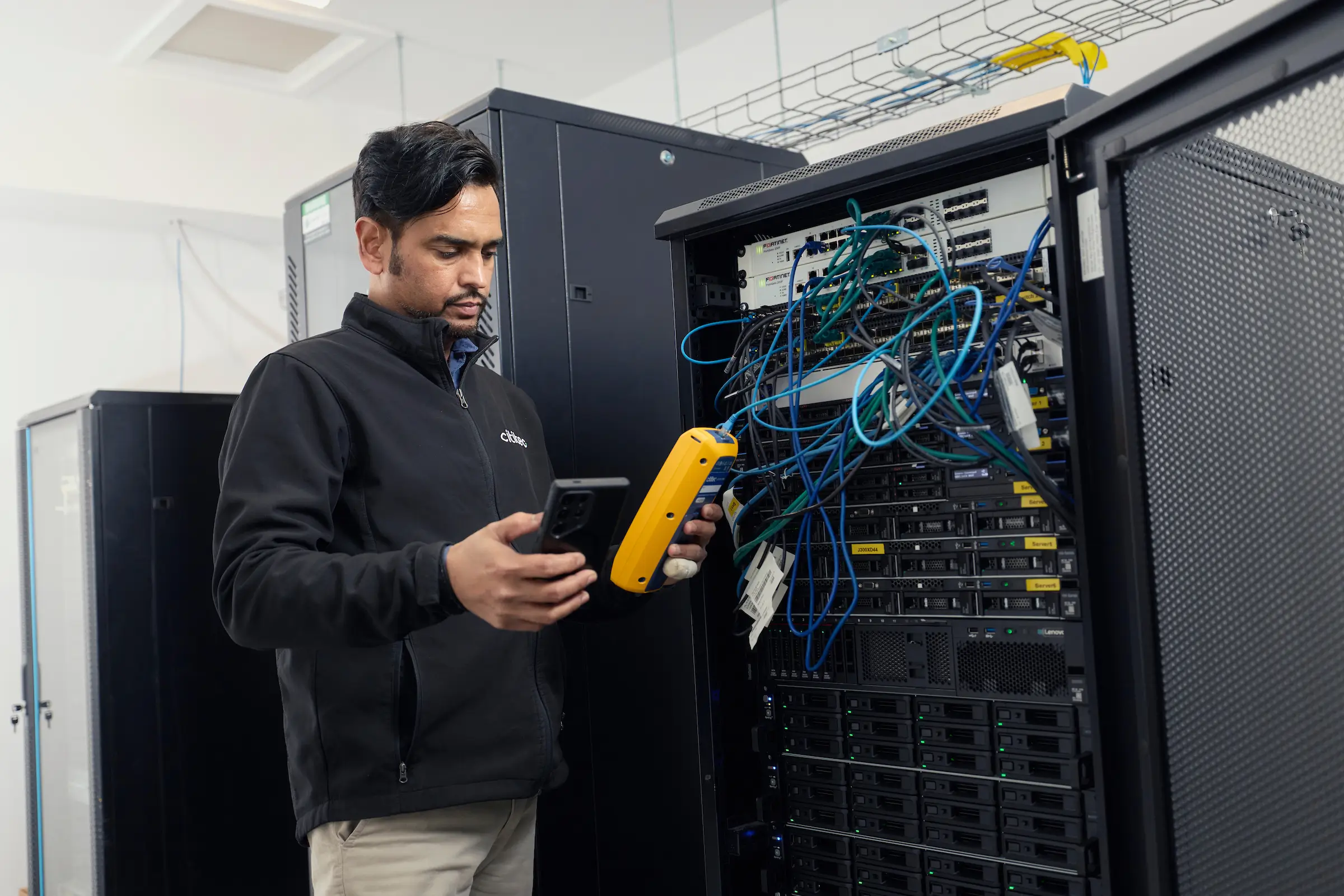A man in a server room stands next to a server rack with tangled cables, holding a device and a smartphone, appearing to check or troubleshoot equipment.