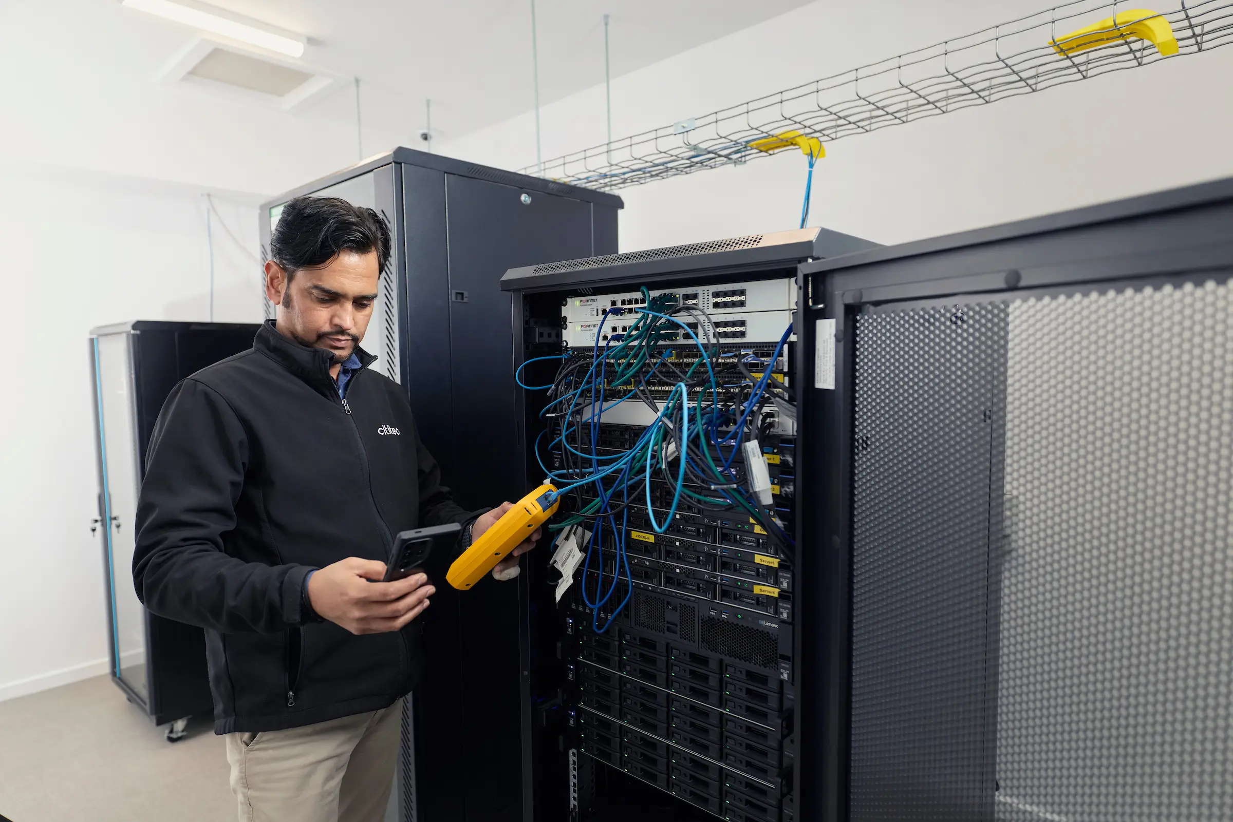 A man in a server room tests network cables connected to a rack-mounted server using diagnostic equipment.