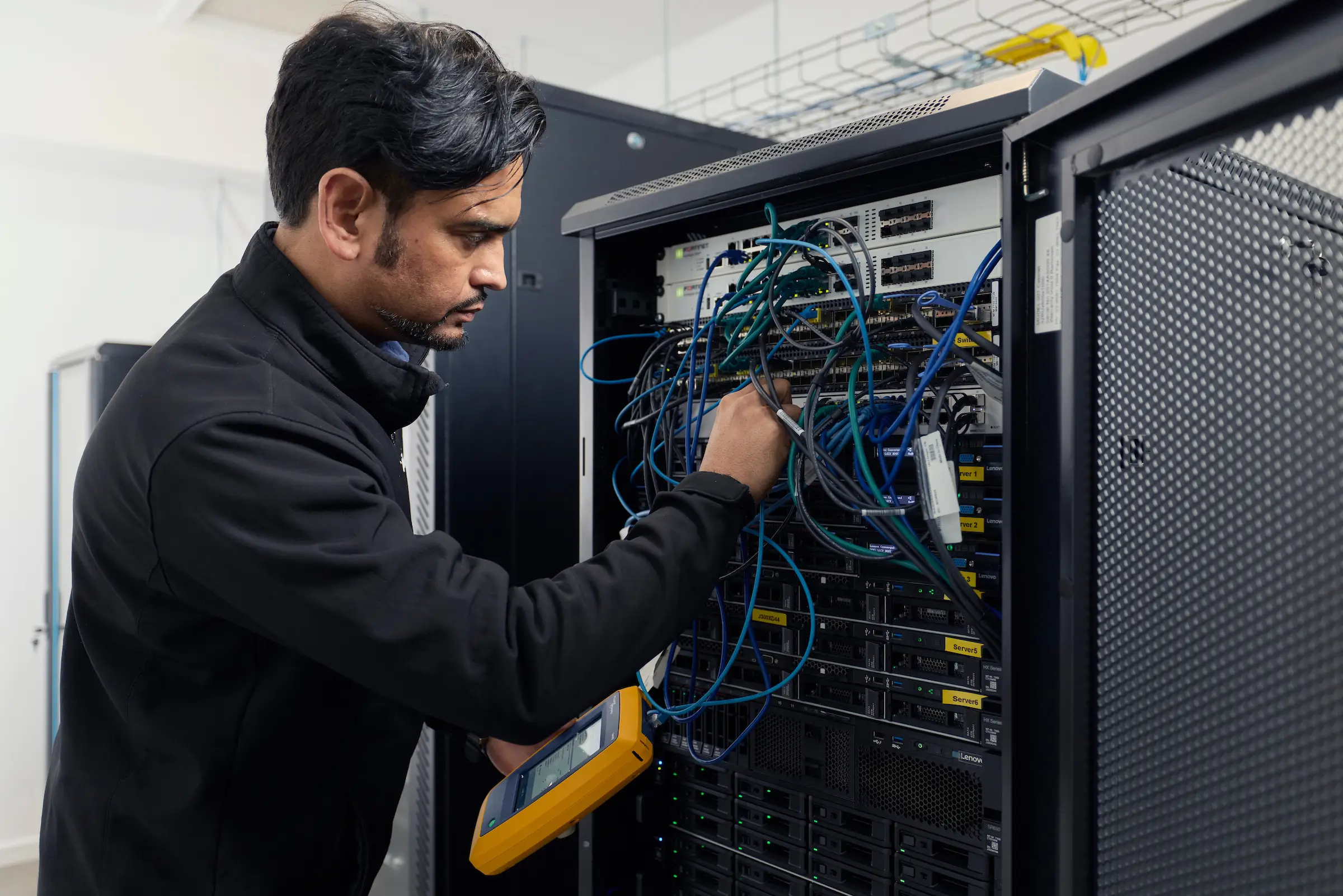 A technician uses a handheld device to test network cables connected to a server rack in a data center.