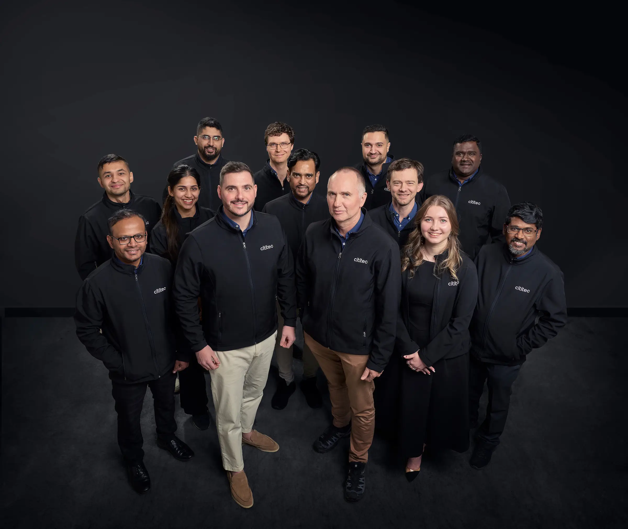 A group of twelve people, wearing matching black jackets with "CEAD" logos, stand together and face the camera against a plain dark background.